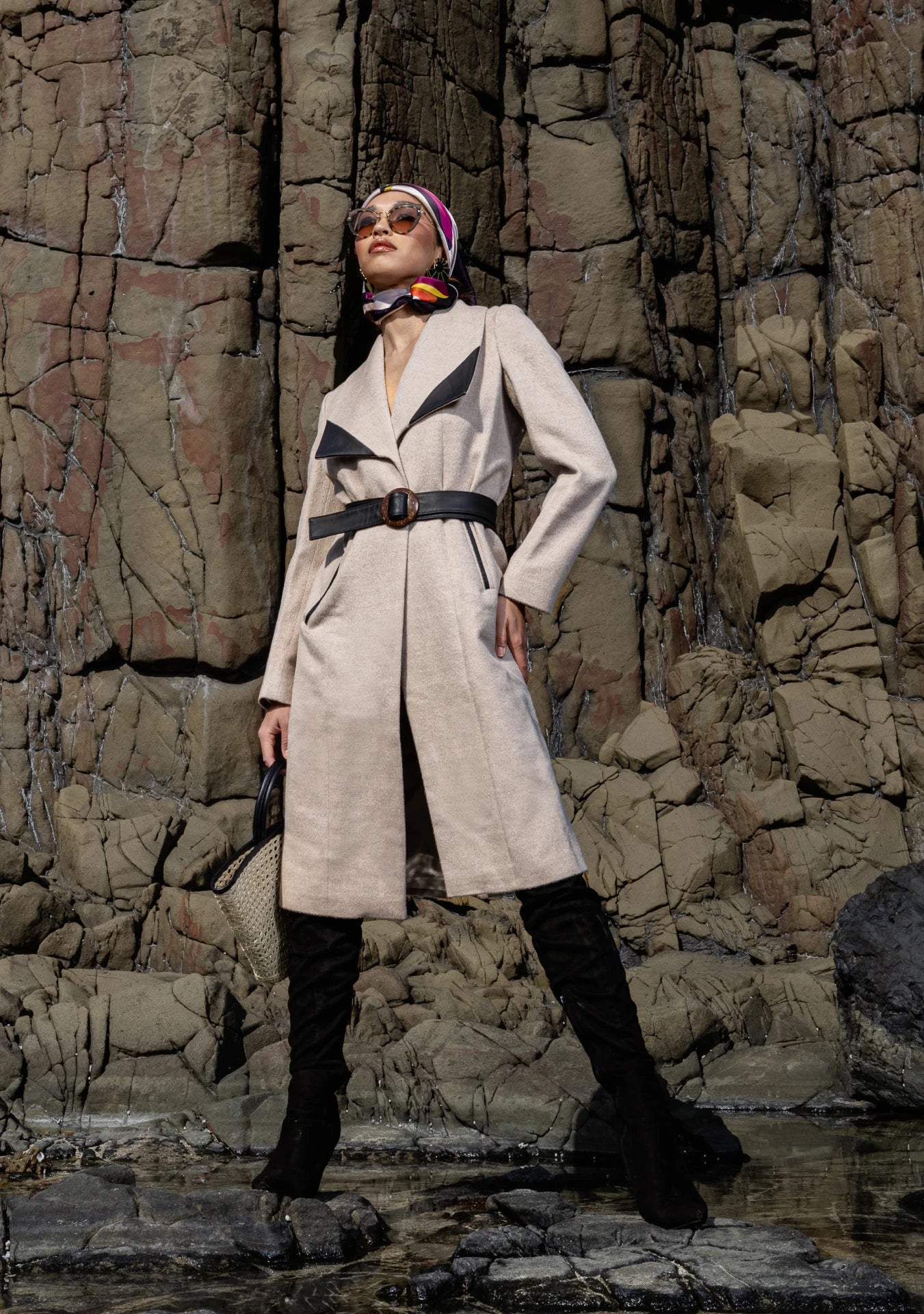 Girl wearing the Mia Belted Taupe Coat with wide lapels and black accents, captured in Madrid, Spain – timeless tailoring by My Paloma.  Paired with a straw bag and a silk head scarf on a rock shore