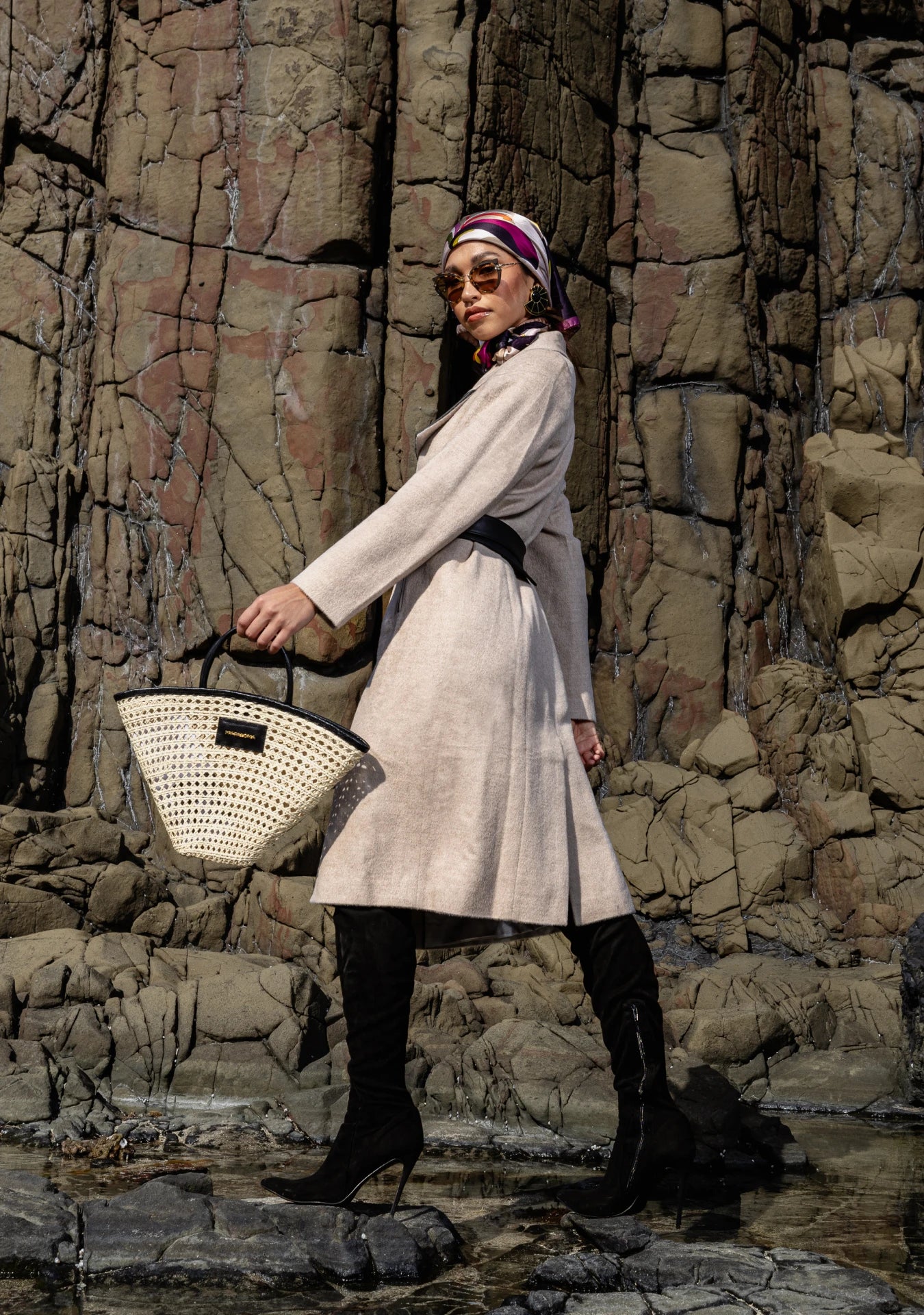 Girl wearing the Mia Belted Taupe Coat with wide lapels and black accents, captured in Madrid, Spain – timeless tailoring by My Paloma.  Paired with a straw bag and a silk head scarf on a rock shore