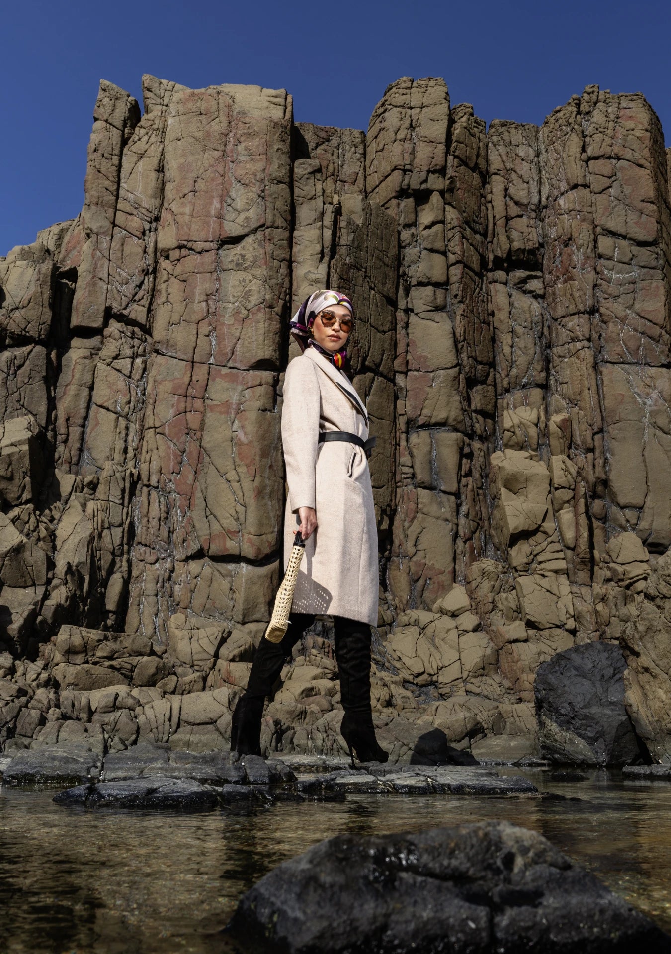 Girl wearing the Mia Belted Taupe Coat with wide lapels and black accents, captured in Madrid, Spain – timeless tailoring by My Paloma.  Paired with a straw bag and a silk head scarf on a rock shore