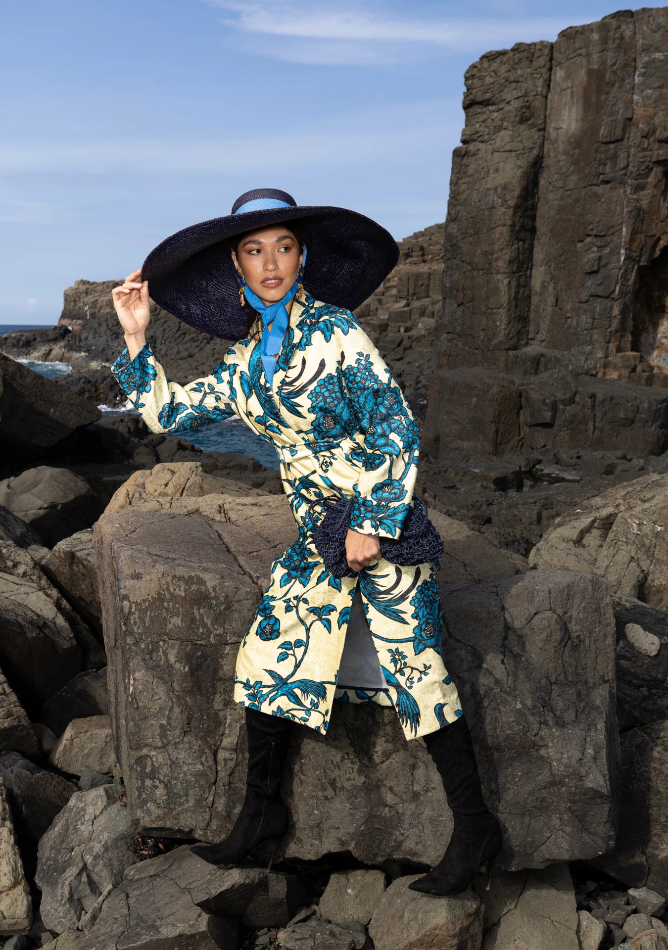 Seated model in the Lady Ibiza Hat in navy, paired with a layered, floral ensemble on dark rock formations—evoking an exotic, high-fashion statement in raw outdoor settings.