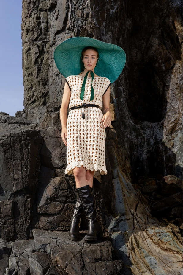 Full view of the model in the Mira crochet dress, highlighting the intricate weave and relaxed drape of the fabric amidst dramatic cliffside surroundings.