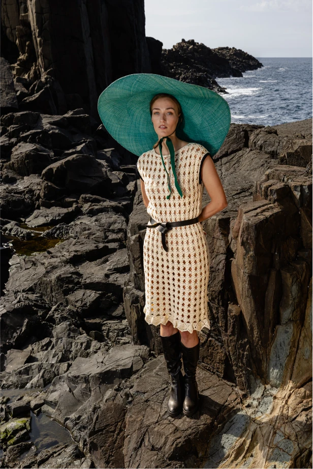 Model turned slightly to the side wearing the Mira dress, capturing the detailed texture and scalloped hemline against a moody, rugged backdrop.