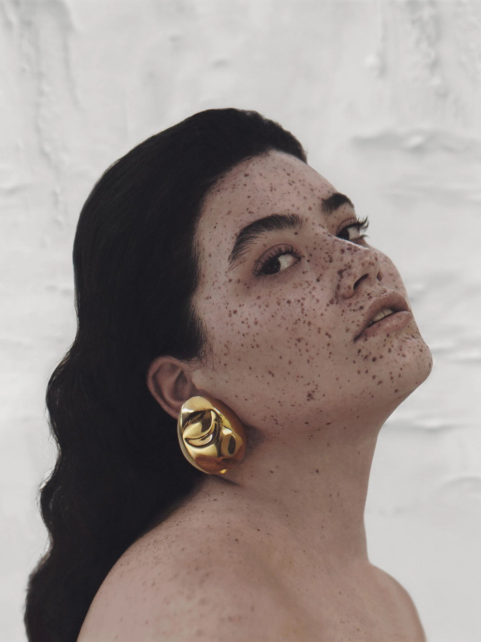 Model with dark, wavy hair wears gold sculptural stud earrings, styled to accentuate their bold and artistic shape.