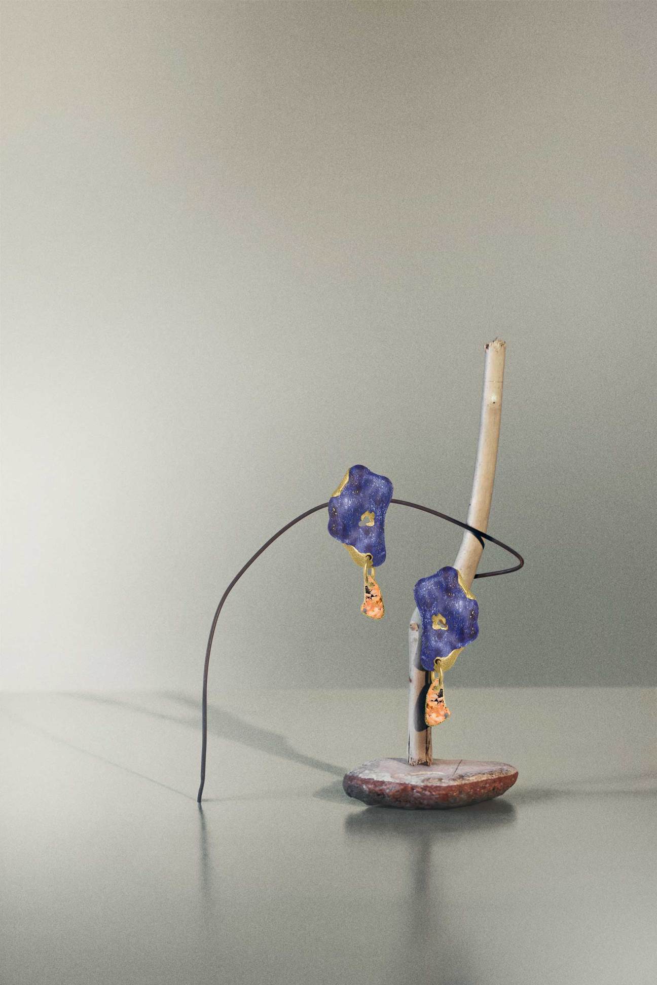 Sculptural gold-plated earrings with organic blue and orange stones displayed on a minimalist wire stand, resembling wearable art.