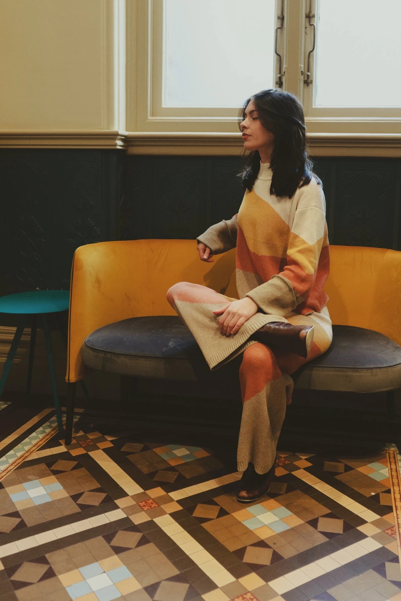 Model sitting cross-legged on a velvet couch in the My Paloma knit set, surrounded by vintage mosaic tiles and natural light, showcasing the outfit's cozy elegance and statement pattern.

